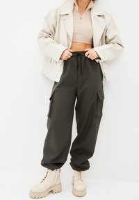 Chaqueta de piel sintética beige, top de punto beige corto, pantalones cargo verde oliva con bolsillos, botas beige gruesas con suelas texturizadas.