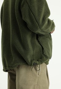 Zielony polarowy sweter o teksturowanej tkaninie, z gładką kieszenią w ciemniejszym odcieniu, regulowanym sznurkiem na dolnym brzegu oraz luźnym kroju.