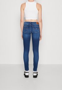 Jeans de denim azul ajustados con una cintura de media altura, dos bolsillos traseros y una etiqueta de marca marrón. Combinados con calcetines blancos y zapatos negros.