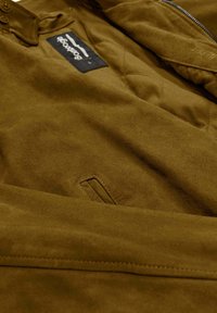 Braune Wildlederjacke mit gequiltem Muster, ausgestattet mit einem Reißverschluss, Logosticker und einer kleinen Tasche. Glatte Oberfläche mit einem weichen Erscheinungsbild.