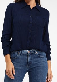 Granatowa koszula z długim rękawem w pionowe paski, zapinana na guziki, z kołnierzykiem i jedną kieszenią na piersi, w zestawie z dżinsami.