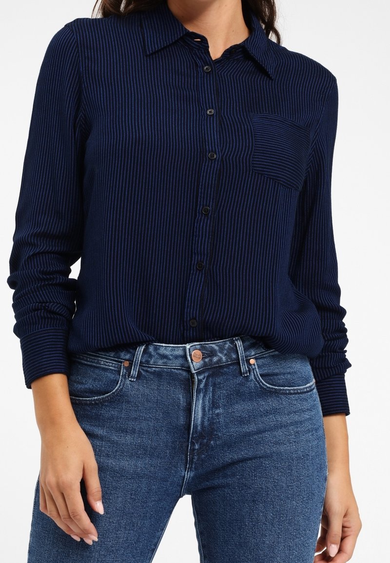 Granatowa koszula z długim rękawem w pionowe paski, zapinana na guziki, z kołnierzykiem i jedną kieszenią na piersi, w zestawie z dżinsami.