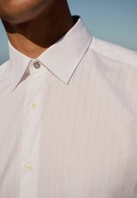 White collared shirt featuring thin vertical stripes, a smooth texture, and a single button detail at the collar with a subtle sheen.