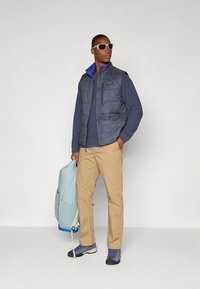 Patagonijos pūkinė liemenė tamsiai pilkos spalvos, dėvima ant tamsiai mėlynos ilgomis rankovėmis marškinėlių, smėlio spalvos kelnių, pilkų batų, laikant šviesiai mėlyną kuprinę. Akiniai nuo saulės dėvimi.