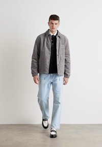 Grå kordfløjljakke med et logo, sort polo-shirt, lys blå jeans og tofarvede sko. Model står foran en ensfarvet baggrund.
