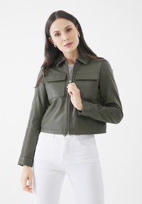 Grün-beige Lederjacke mit Kragen, zwei vorderen Taschen und silbernem Reißverschluss. Getragen über einem hellgrauen Oberteil und weißen Jeans.