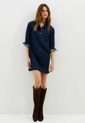 Mujer con un vestido de mezclilla azul oscuro con las mangas remangadas y botas marrones hasta la rodilla, de pie frente a un fondo claro y liso.