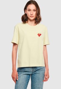 Woman wearing a pale yellow t-shirt with a small red cherry graphic on the left chest, paired with blue jeans, standing against a plain background.