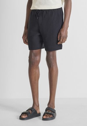 Uomo che indossa pantaloncini neri con coulisse in tessuto strutturato e sandali neri aperti in punta, in piedi davanti a uno sfondo chiaro e uniforme.