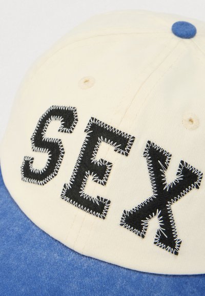 Șapcă de baseball cu partea din față din bumbac crem și vizieră din denim albastru. Prezintă litere brodate negre „SEX” pe față, cu cusături decorative albe.
