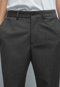 Pantaloni tailored grigio scuro con una texture liscia, chiusura con bottone e tasche laterali. Presentano una vestibilità classica e un sottile motivo a righe diagonali.