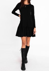 Schwarzes langärmeliges Kleid mit einem ausgestellten, gefransten Saum, aus weichem Stoff. Kombiniert mit hohen schwarzen Schnürstiefeln und einer dezenten Kreuzkette.