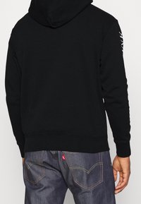 Hombre vistiendo una sudadera negra con texto blanco en la manga y jeans oscuros con costuras visibles en los bolsillos traseros.