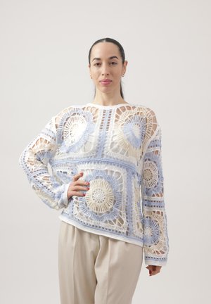 Mujer con cabello oscuro recogido lleva un suéter blanco de crochet con patrones circulares en azul y beige, y pantalones beige, posando con la mano en la cadera.