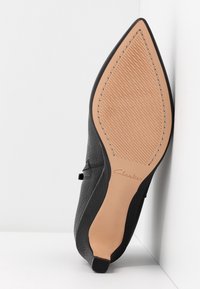 Chaussure noire à bout pointu montrant une semelle beige texturée avec de fines lignes diagonales et l'impression du nom de la marque Clarks.