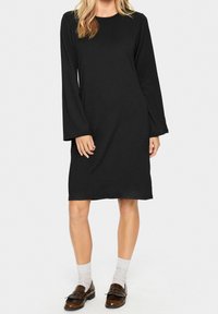 Svart långärmad klänning med rund ringning, avslappnad passform och knä-lång nederkant. Bärs med grå strumpor och loafers. Slät textur.