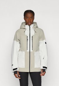 Kevyt, vedenpitävä takki beige ja valkoinen, jossa on huppu, etuvetoketju, useita taskuja ja mustia yksityiskohtia.