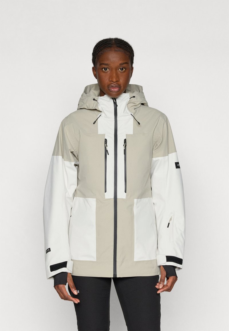 Kevyt, vedenpitävä takki beige ja valkoinen, jossa on huppu, etuvetoketju, useita taskuja ja mustia yksityiskohtia.