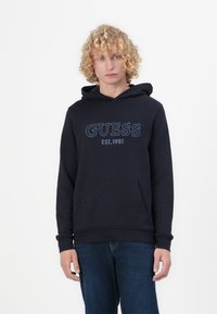 Hoodie blu navy con tasca a canguro, presenta la scritta "GUESS EST. 1981" in cuciture azzurre, realizzato in morbido tessuto misto cotone.