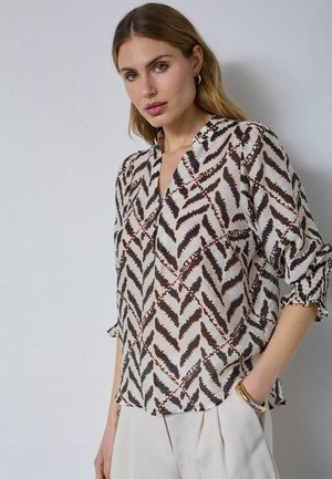 Femme portant un chemisier à manches longues à motifs zigzag noirs et marron, associé à un pantalon clair et des boucles d’oreilles créoles dorées.