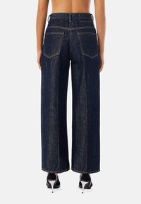 Tmavě prané džínové kalhoty se širokými nohavicemi, zadními kapsami a výraznými švy, stylizované s vysokými podpatky.