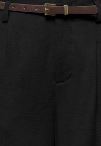 Ceinture en cuir marron avec boucle dorée attachée sur un pantalon en tissu noir texturé avec une poche latérale visible.