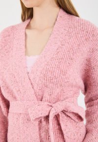 Rozā adīta kardigāna ar ribotu tekstūru, platām atlokām un sasietu vidukļa jostu. Audums satur nelielus punktiņus, kas piešķir papildu detaļas.