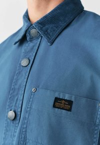 Bl�å skjorte med knapper og en struktureret fløjlskrave, brystlomme og et lille brandmærke. Har trykknapper og en glat stoffinish.