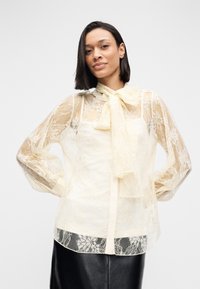 Δαντελένιο μπλουζάκι σε κρεμ απόχρωση με διαφανείς μακριές μανσέτες, γιακά με φιόγκο και ανθικό σχέδιο στις μανσέτες και στο τελείωμα, με στερεή επένδυση από κάτω.