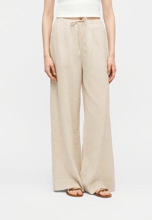Donna che indossa pantaloni beige a gamba larga con coulisse in vita e top bianco corto, in piedi con le mani rilassate lungo i fianchi, sandali marroni visibili.