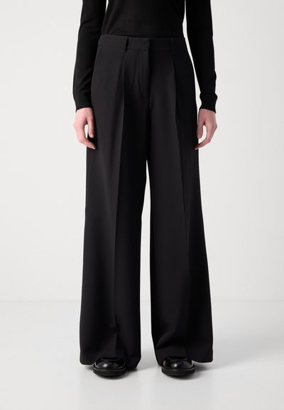 Pantalons larges noirs en tissu lisse avec des plis devant, offrant une coupe ajustée et une taille haute. Chaussures en cuir noires incluses.