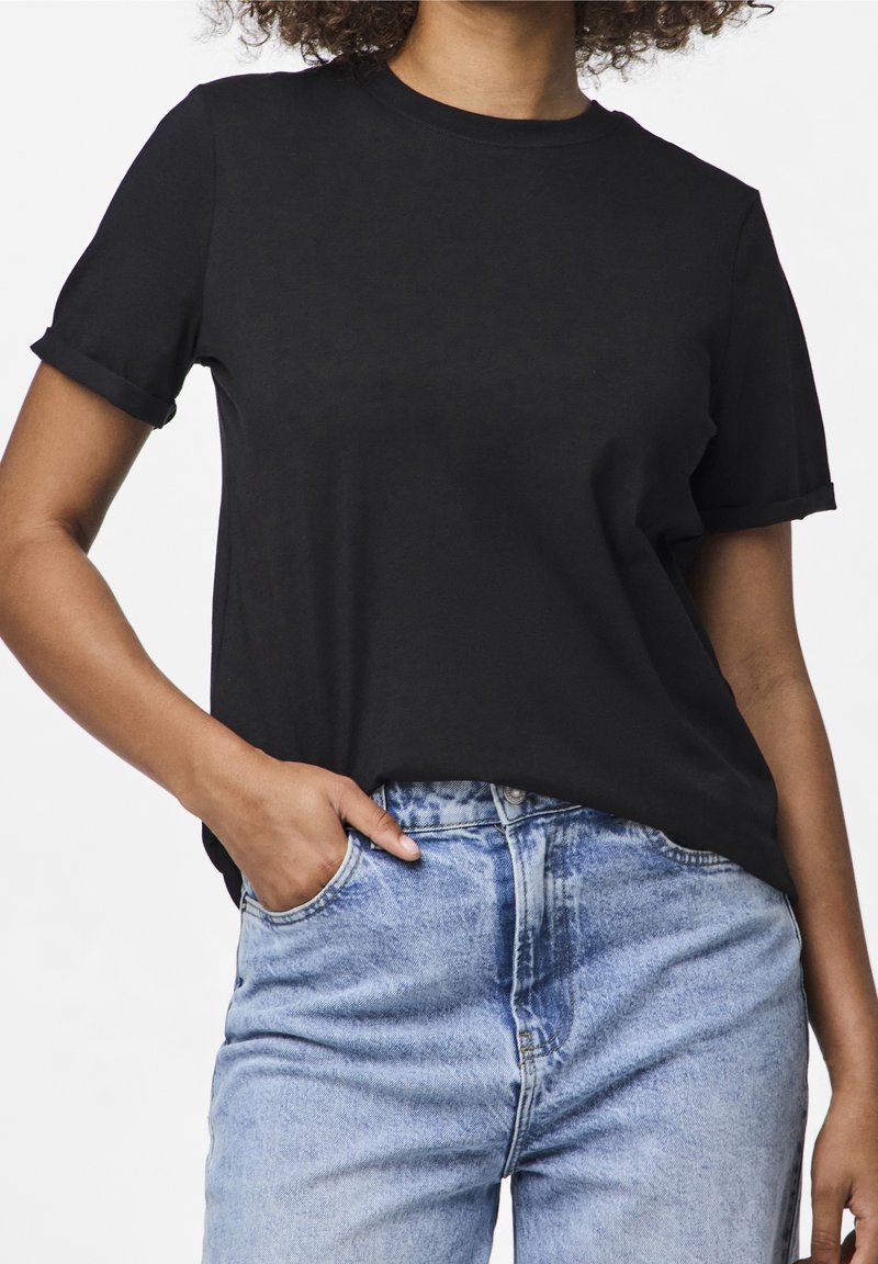 Femme portant un T-shirt noir uni à manches courtes, légèrement rentré dans un jean taille haute bleu clair, une main dans la poche.