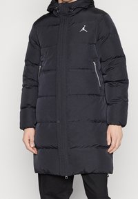 Persoon in een zwart gewatteerd langjack met een capuchon en ritssluitingen aan de zijkanten, voorzien van een wit Jumpman-logo op de borst.