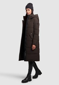 Brun polstret jakke med hætte, lang længde, sidelommer og sorte lynlåse, parret med en mørk hue og tætsiddende sorte leggings og støvler.