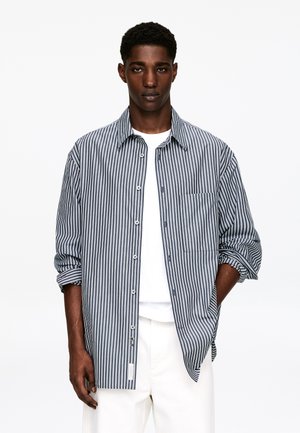 Koszula na guziki w paski w kolorze granatowym i białym, z pojedynczą kieszenią na piersi, podwiniętymi rękawami i luźnym krojem, noszona na białym t-shircie.