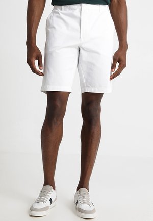 Valkoiset puuvillashortsit suoralla leikkauksella, joissa on litteä etuosa, sivutaskut ja sileä pinta. Yhdistetty valkoisiin tennareihin ja vihreään toppiin.