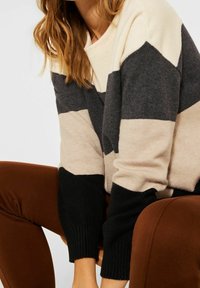 Kvinna i en färgblockerad tröja i beige, grå och svart med rostfärgade byxor, sittande med armbågarna vilande på knäna.