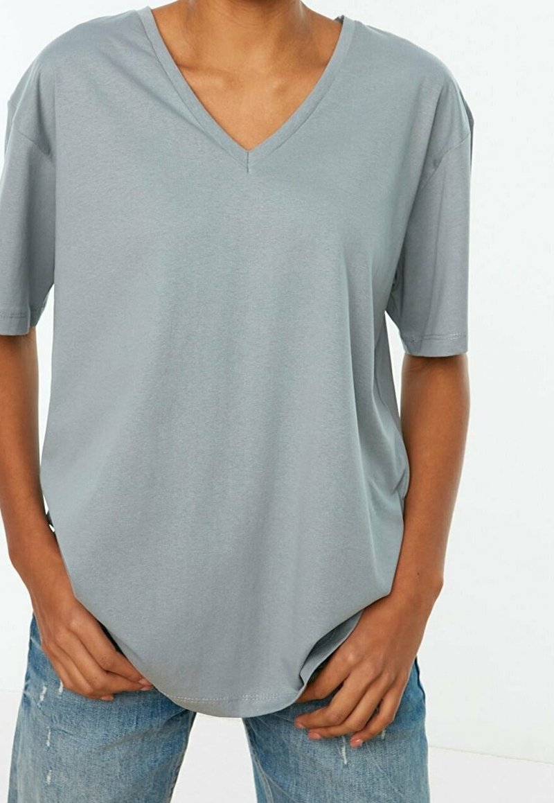 T-shirt gris clair à manches courtes en V, fabriqué en tissu doux, avec une coupe décontractée et un ourlet légèrement arrondi. Porté avec un jean bleu déchiré.