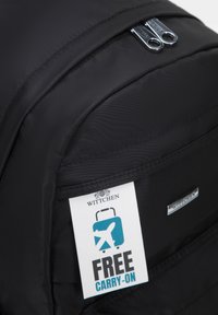 Mochila negra de nailon con dos cremalleras plateadas, que presenta una etiqueta blanca con la inscripción "CARRI-ON GRATIS" y un sutil detalle de logotipo.