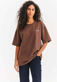 Brązowy oversize t-shirt wykonany z bawełny z krótkimi rękawami. Na lewej piersi znajduje się mała biała grafika rzęs. Połączony z ciemnymi dżinsami.