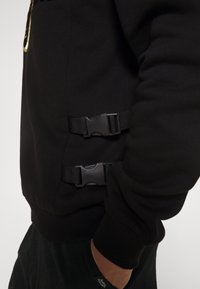 Felpa nera con superficie testurizzata, dotata di due chiusure laterali e polsini a costine. Il design include una vestibilità comoda, con una silhouette regolabile.