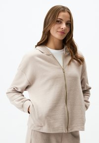 Felpa con zip beige realizzata in un tessuto morbido, dotata di cappuccio, maniche lunghe e zip dorata. Include tasche laterali e vestibilità rilassata.