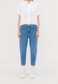 Vit kortärmad skjorta med knappad krage, i kombination med ljusblå högmidjade jeans och vita sneakers, stående mot en neutral bakgrund.