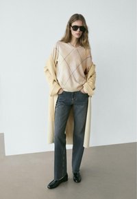 Maglione beige con motivo e scollo a V, jeans grigi a gamba dritta e mocassini neri. Sovrapposto a un lungo cappotto giallo pallido.
