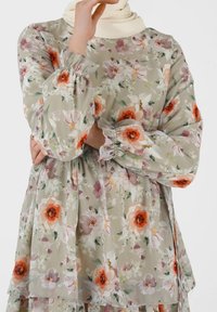 Blomstret tunika i lys grøn, med puffede ærmer med elastiske manchetter, lagdelt design og mønstre af orange og lilla blomster.