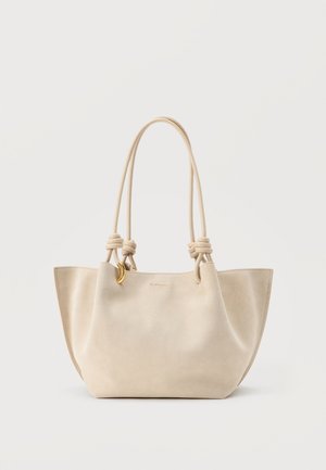 Borsa tote in morbida pelle beige con lunghi manici doppi annodati e piccolo ciondolo a mezzaluna dorato su sfondo semplice.