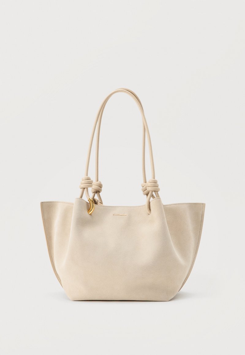 Sac cabas en cuir souple beige avec longues poignées doubles nouées et petit charme en forme de croissant doré sur fond uni.