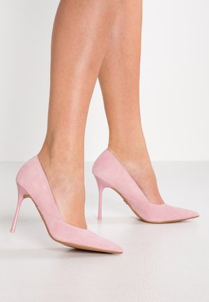 Kvinde iført spidse pink ruskindsstiletter med slanke stiletter, stående på en glat hvid gulv.