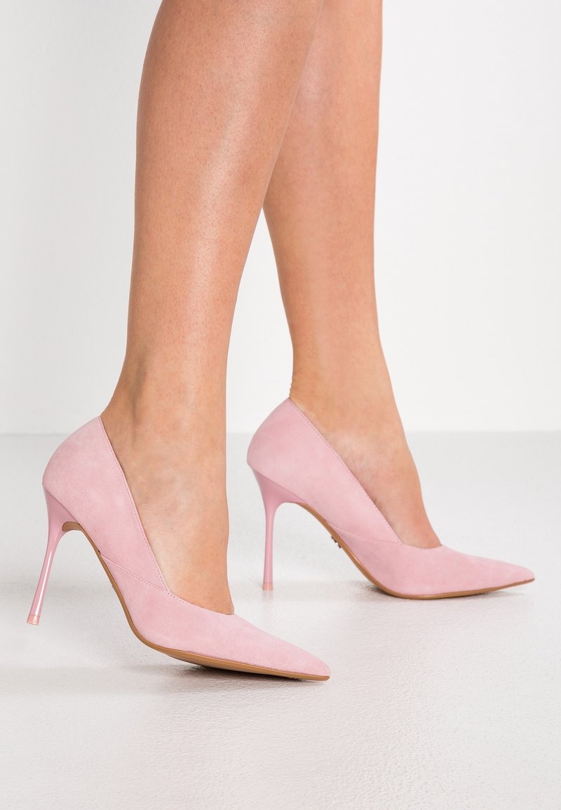 Femme portant des escarpins en daim rose à bout pointu avec des talons aiguilles fins, debout sur un sol blanc lisse.