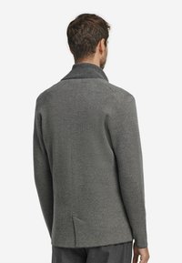 Grå stickad jacka med sjalkrage, textilerad tyg och detalj med slits bak. Har långa ärmar och rena sömmar.